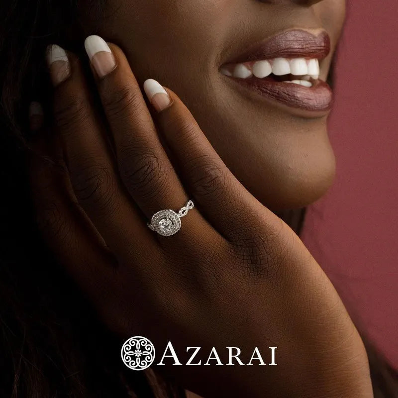 A person smiles while touching their face, showcasing a stunning Astrid sterling silver engagement ring on their finger. The logo "AZARAI" is visible at the bottom.