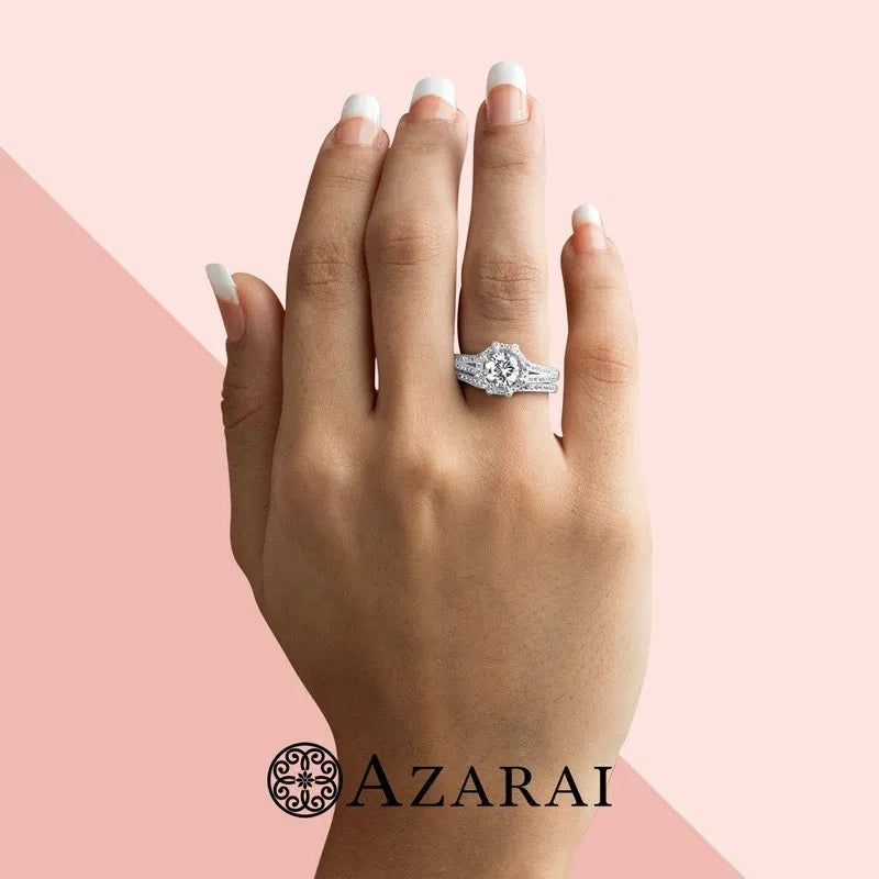 A hand with manicured nails elegantly presents the Britney sterling silver bridal set ON CLEARANCE against a light pink and peach backdrop. The Azarai logo is subtly placed below, while the sterling silver band shines in sophistication, completing the picture-perfect look.