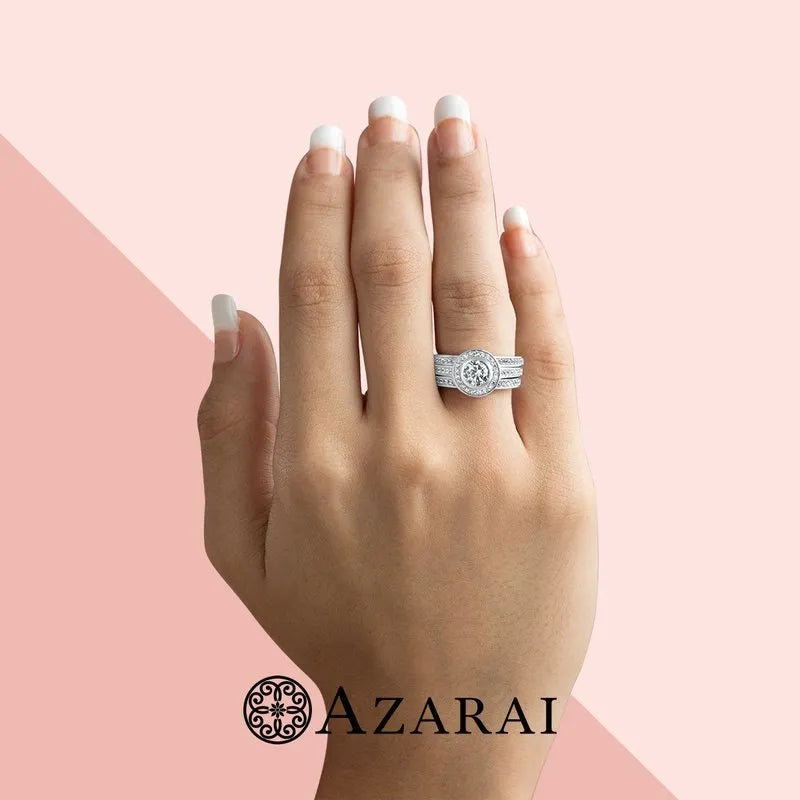 A hand with manicured nails displays the Chamise sterling silver bridal set ON CLEARANCE, featuring a dazzling large diamond ring alongside a smaller band, both shimmering in sterling silver against a pink and light pink backdrop. The word "AZARAI" is gracefully featured at the bottom.