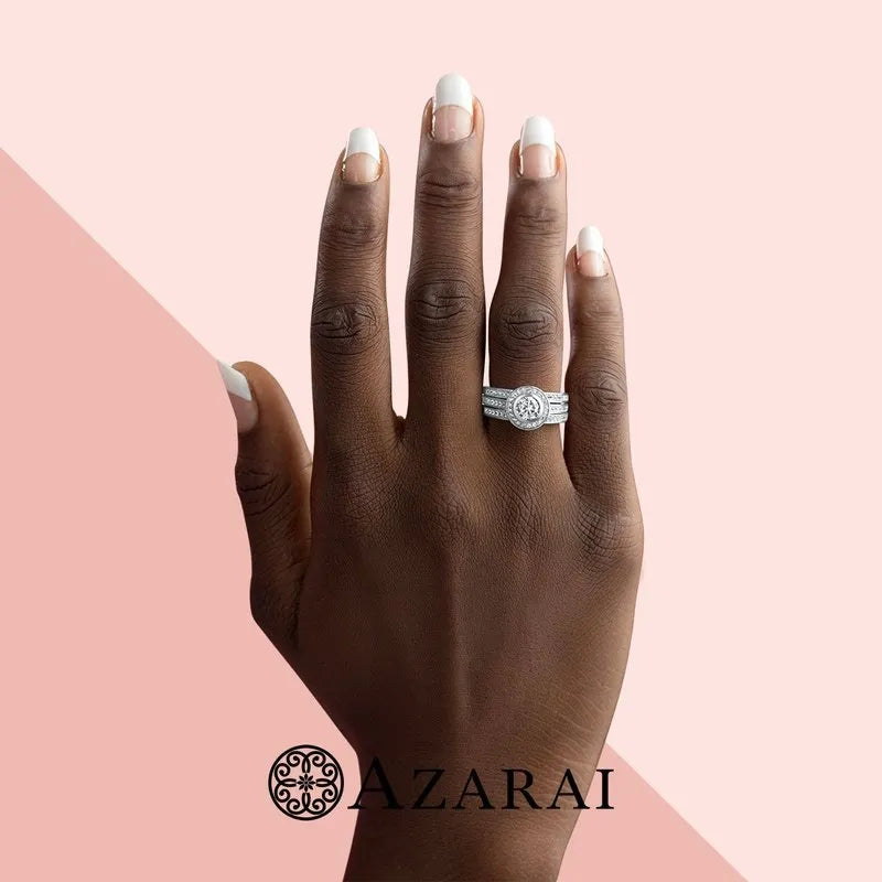 A hand with a French manicure displays a sparkling diamond ring from the Chamise sterling silver bridal set, currently on clearance, against a pink and beige backdrop featuring the Azarai logo.