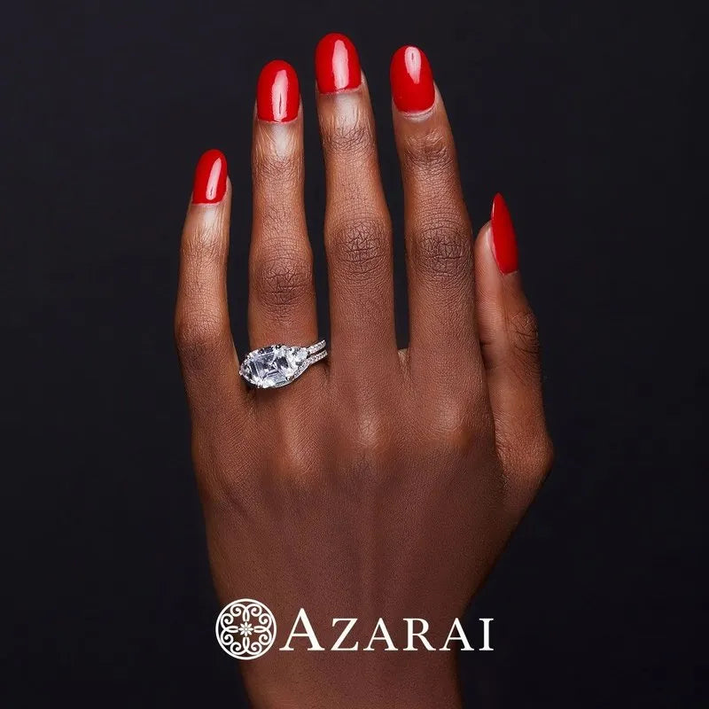 A hand with red nail polish showcases a stunning diamond ring, part of the exquisite Guinevere sterling silver bridal set, against a black background featuring the Azarai logo.