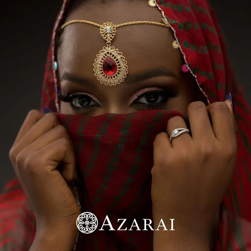 A woman covers her lower face with a red patterned veil, accentuating the gold and red headpiece she wears. The Paloma sterling silver engagement ring on her finger complements her mysterious allure, with the word "Azarai" elegantly displayed at the bottom.