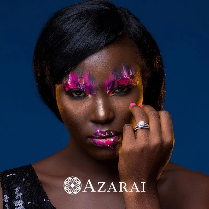 A person with artistic pink and purple makeup poses against a blue background, elegantly displaying an Eva sterling silver bridal set from the clearance section on their finger. The word "AZARAI" is visible at the bottom.