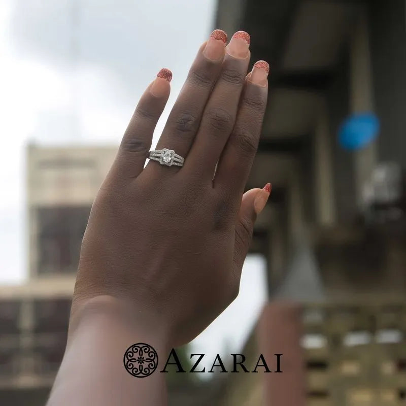 A hand with polished nails elegantly displays a dazzling diamond engagement ring against a blurred urban backdrop. The word "Azarai" is prominently featured at the bottom, hinting at the allure of the Esmee sterling silver engagement ring on clearance.