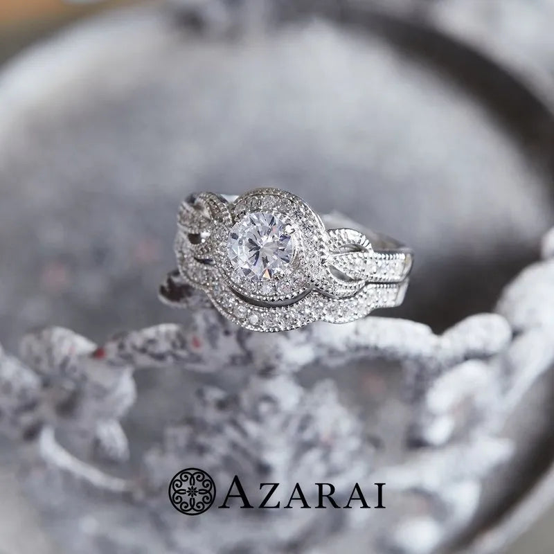 A captivating Louisa sterling silver bridal set ON CLEARANCE, adorned with intricate detailing and sparkling diamonds, is showcased on an ornate metal surface. The Azarai logo shines brightly at the base, drawing attention and enhancing the allure of this remarkable piece.